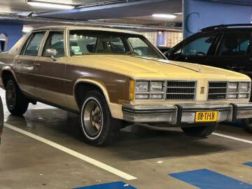 Een beige en bruine vintage Oldsmobile Delta 88 staat geparkeerd op een overdekte parkeerplaats, geflankeerd door een zilveren auto aan de linkerkant en een zwarte SUV aan de rechterkant. De auto heeft een geel kenteken met de tekst "08-TL-H".