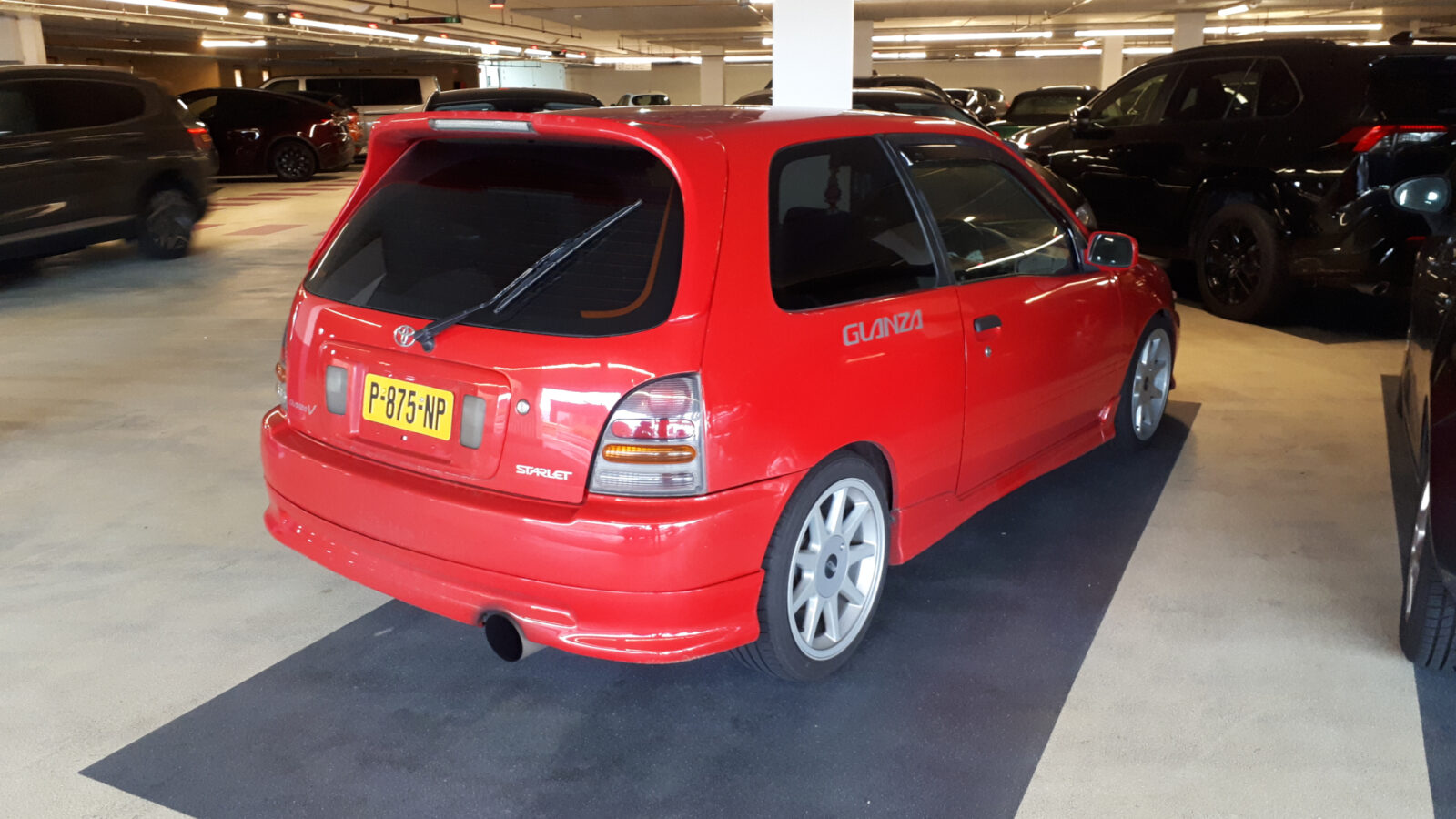 In een ondergrondse parkeergarage staat een rode Toyota Starlet hatchback met spoiler geparkeerd. De auto heeft een kentekenplaat met de tekst "P? 75 HIP" en een "GLANZA"-sticker op de zijkant.