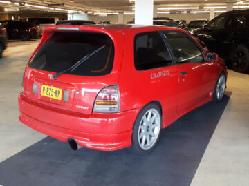 In een ondergrondse parkeergarage staat een rode Toyota Starlet hatchback met spoiler geparkeerd. De auto heeft een kentekenplaat met de tekst "P? 75 HIP" en een "GLANZA"-sticker op de zijkant.