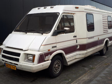Een witte Winnebago LeSharo met kastanjebruine strepen staat geparkeerd in een straat naast een gebouw. Het voertuig vertoont tekenen van slijtage en roest. Op de voorzijde bevindt zich een Europees kenteken.