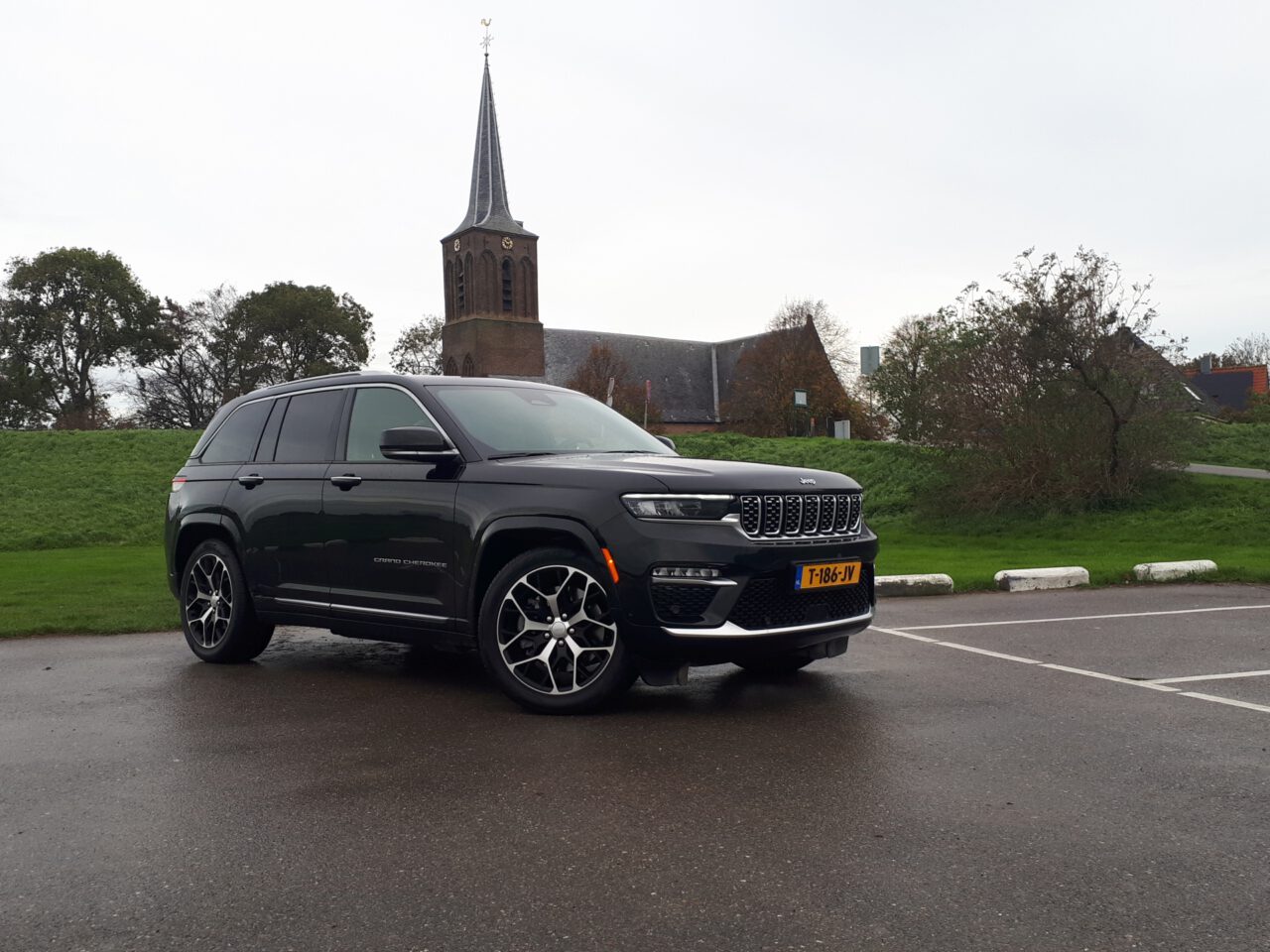 Een zwarte Jeep Grand Cherokee staat geparkeerd op een natte stoep voor een grasveld met een kerk met een hoge toren en enkele bomen op de achtergrond.