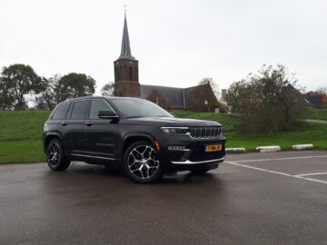 Een zwarte Jeep Grand Cherokee staat geparkeerd op een natte stoep voor een grasveld met een kerk met een hoge toren en enkele bomen op de achtergrond.