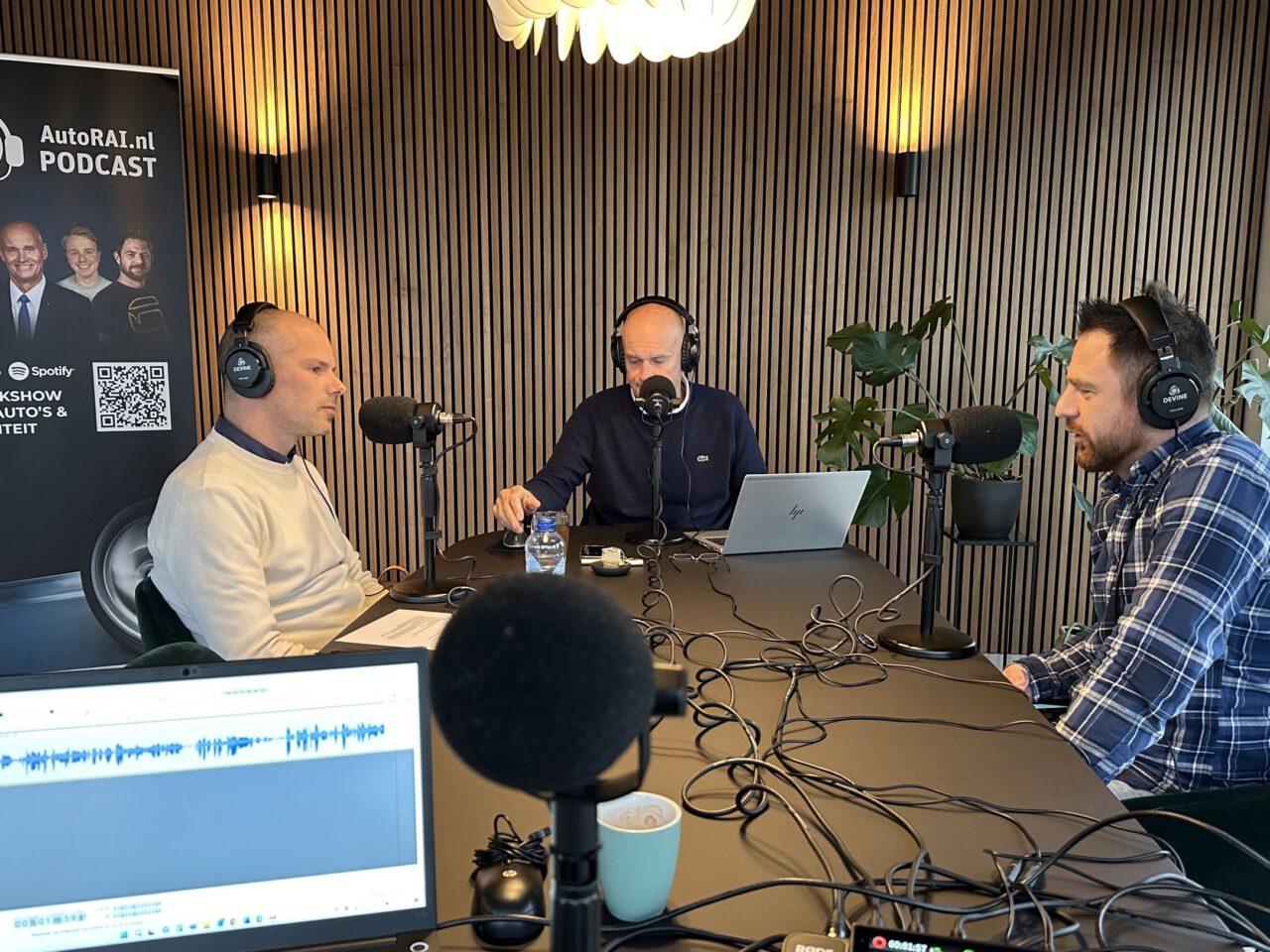 Drie mannen zitten aan een tafel met microfoons en zijn bezig met een AutoRAI Podcast-opname. Een banner op de achtergrond toont "AutoRAI.nl PODCAST" en aanvullende tekst.