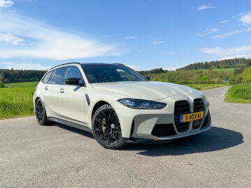 Een witte BMW M3 Touring met zwart dak en donkere wielen staat geparkeerd op een verharde weg in een schilderachtige, landelijke omgeving.