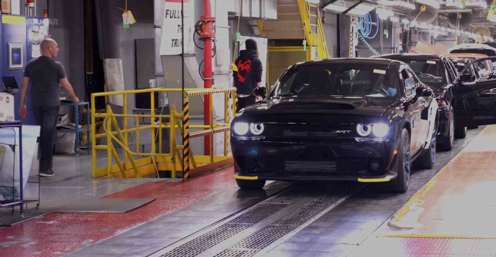 Een strakke zwarte Dodge Challenger SRT glijdt langs een lopende band in de fabriek, met toegewijde werknemers in de buurt.