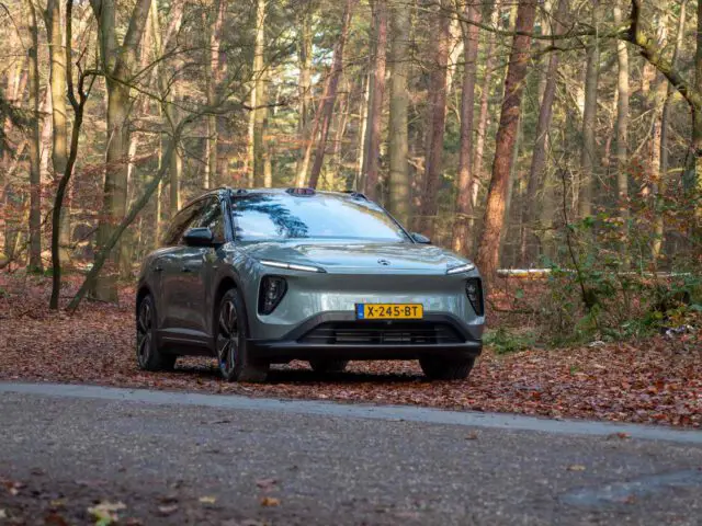 Een zilveren Nio EL6 elektrische auto staat geparkeerd aan de kant van een bosweg bedekt met gevallen bladeren. De bomen op de achtergrond hebben schaars blad, wat duidt op de herfst.