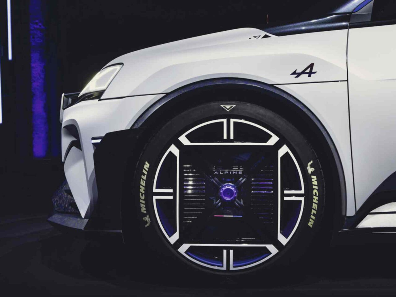 Close-up van het linkervoorwiel van een witte Alpine A290, met een uniek, futuristisch wielontwerp met verlichte elementen en Michelin-logo op de band. De auto beschikt over een sportbadge en een gestileerd logo op het spatbord.
