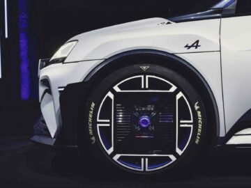 Close-up van het linkervoorwiel van een witte Alpine A290, met een uniek, futuristisch wielontwerp met verlichte elementen en Michelin-logo op de band. De auto beschikt over een sportbadge en een gestileerd logo op het spatbord.