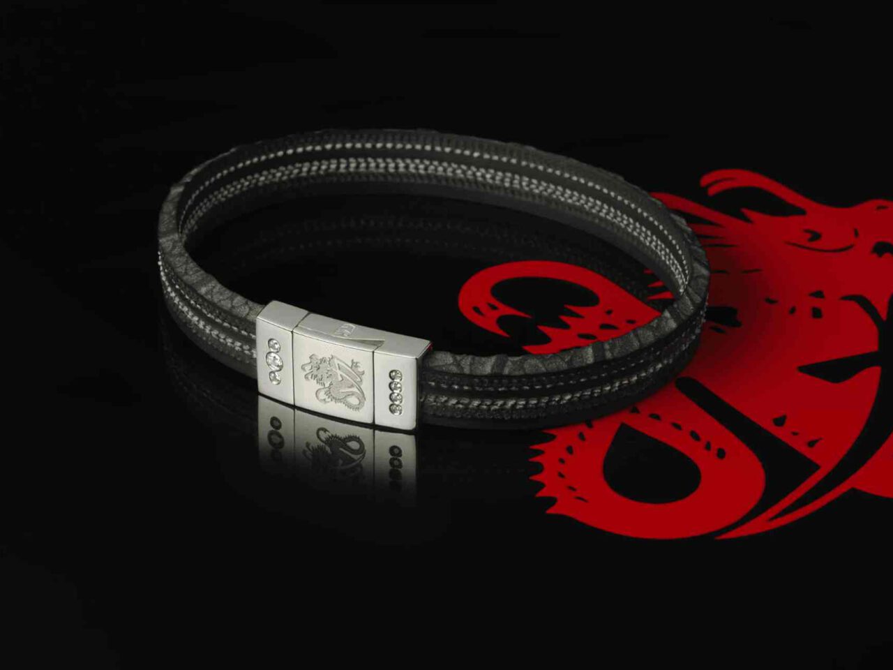 Een zwarte leren armband met een zilveren sluiting en een drakenembleem wordt weergegeven tegen een zwarte achtergrond die doet denken aan de iconische rode drakenafbeelding van Schumacher.