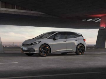 Een slanke zilveren Cupra elektrische hatchback staat geparkeerd in een stedelijke omgeving onder een betonnen structuur, met het stadsbeeld zichtbaar op de achtergrond.