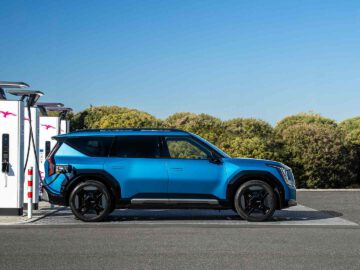 Een blauwe elektrische SUV van Kia staat geparkeerd bij een laadstation, aangesloten op een oplader, in een buitenomgeving met groene struiken en een helderblauwe lucht.