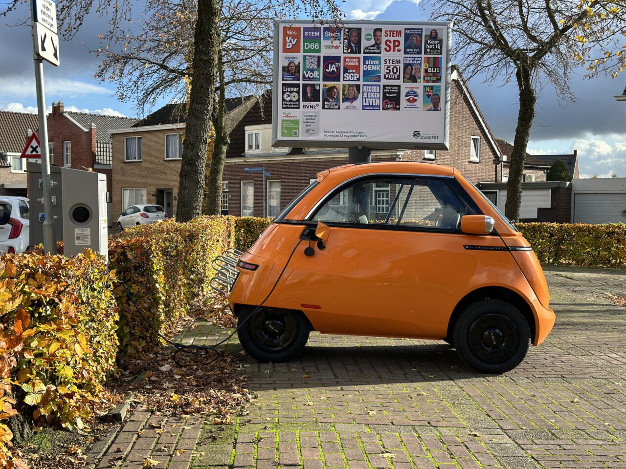 Een kleine oranje elektrische Microlino-auto laadt op bij een openbaar station in een woonwijk, met op de achtergrond een reclamebord met verschillende politieke advertenties.