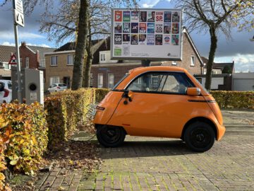 Een kleine oranje elektrische Microlino-auto laadt op bij een openbaar station in een woonwijk, met op de achtergrond een reclamebord met verschillende politieke advertenties.