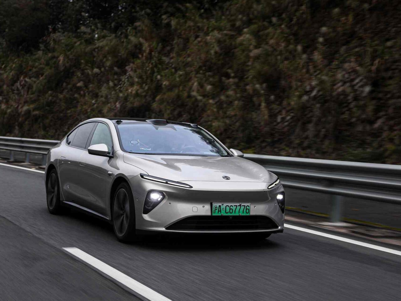 Een zilveren Nio elektrische sedan rijdt op een snelweg met rechts een metalen vangrail en bomen op de achtergrond. De auto heeft een groen kenteken.