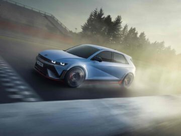 Een blauwe IONIQ 5 N met rode accenten snelt over het circuit, waarachter stof opwaait, met bomen en een wazige tribune op de achtergrond.