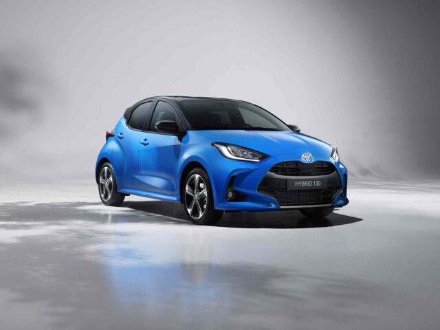 Een blauwe Toyota Yaris hybride compacte auto staat geparkeerd op een reflecterend oppervlak tegen een grijze achtergrond. De auto heeft een modern ontwerp met strakke koplampen en lichtmetalen velgen, wat de eigentijdse uitstraling onderstreept.