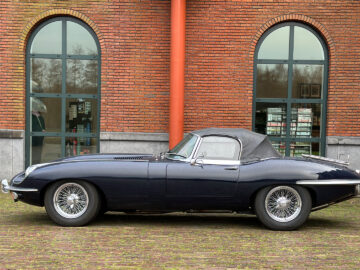 Een zwarte vintage Jaguar E-Type cabriolet staat geparkeerd voor een bakstenen gebouw met hoge boogramen.