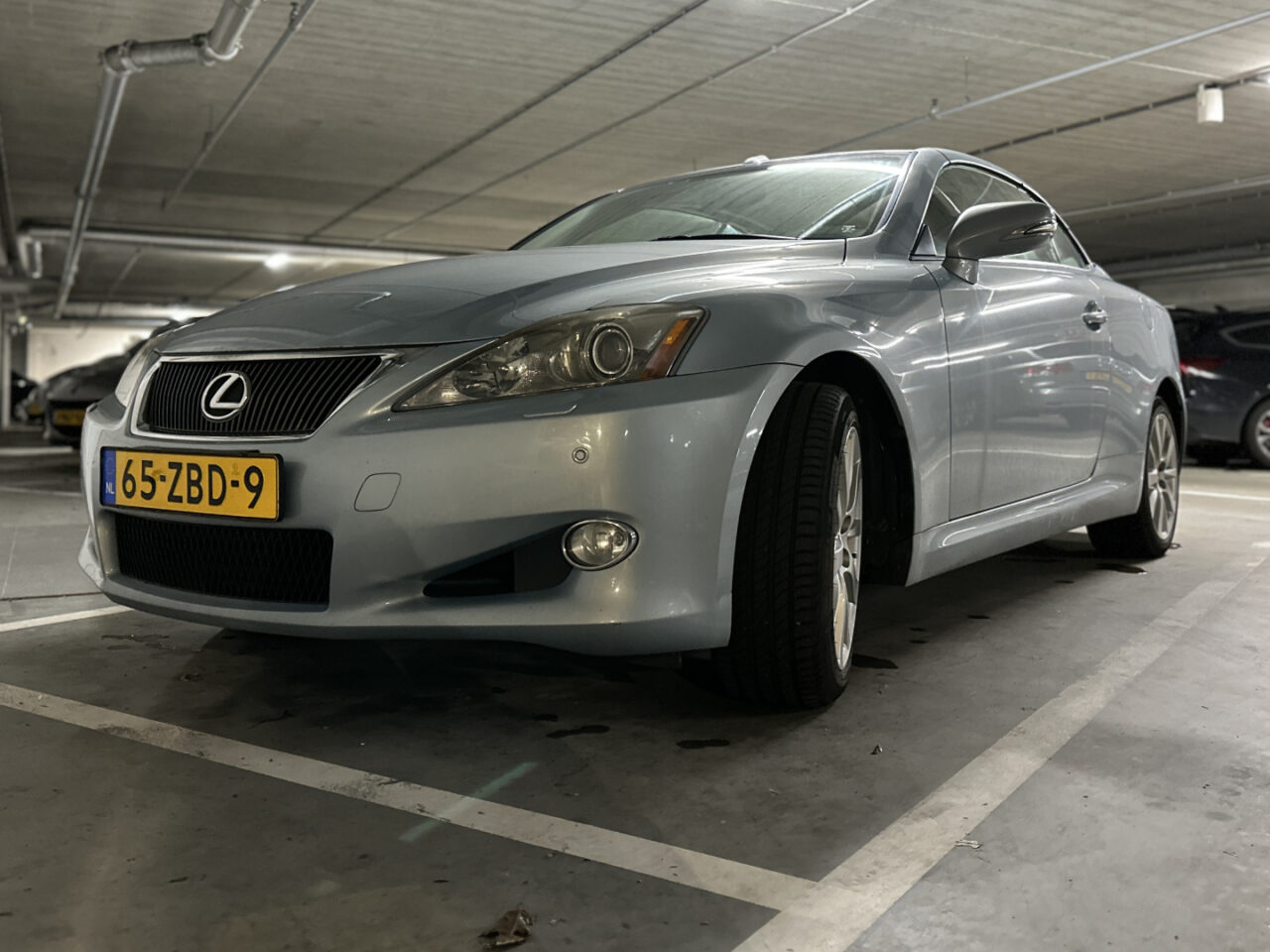 Een zilveren Lexus IS sedan staat geparkeerd in een ondergrondse parkeergarage. De auto heeft een Nederlands kenteken met de vermelding 65-ZBD-9.