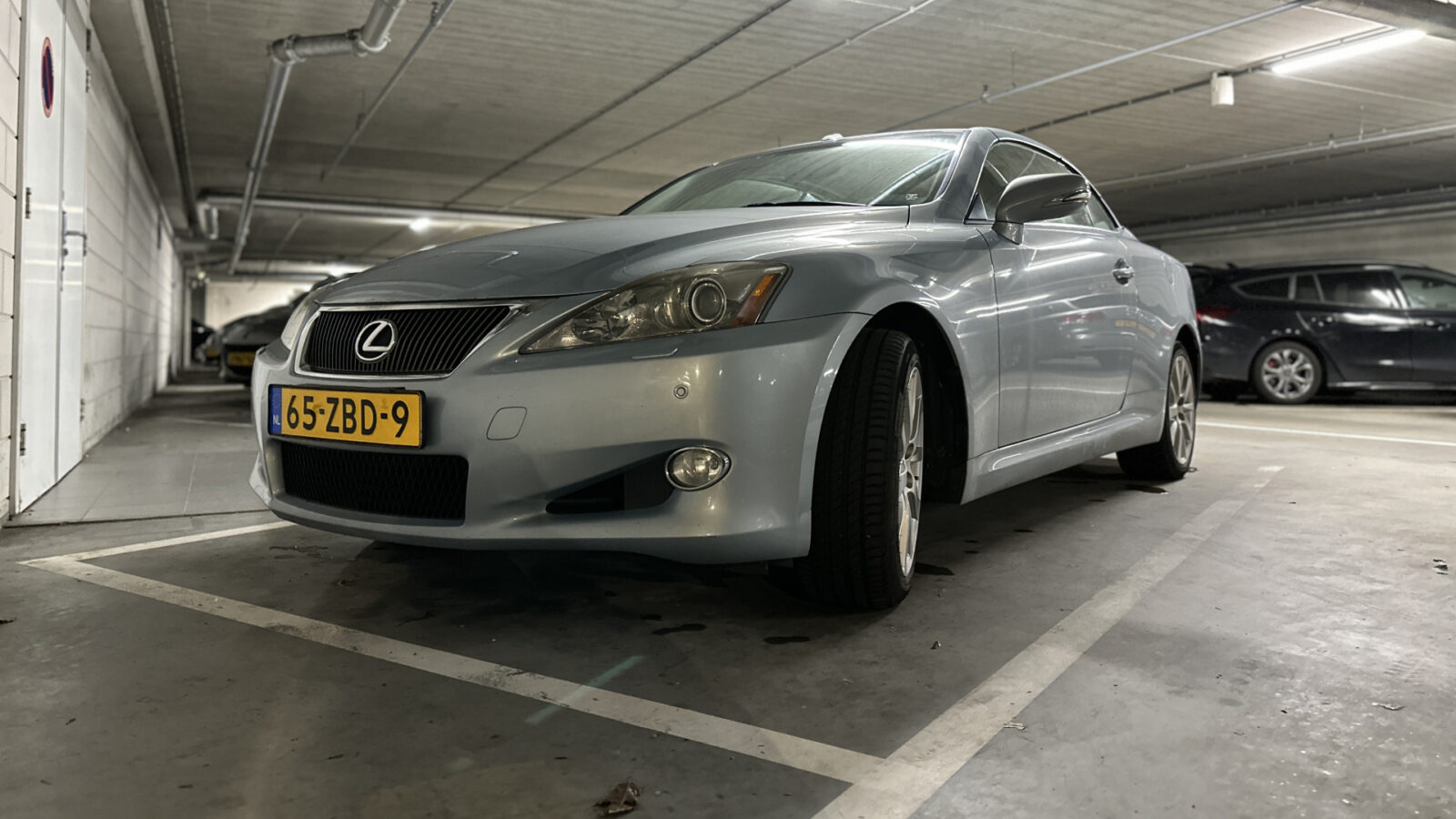 Een zilveren Lexus IS sedan staat geparkeerd in een ondergrondse parkeergarage. De auto heeft een Nederlands kenteken met de vermelding 65-ZBD-9.