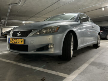 Een zilveren Lexus IS sedan staat geparkeerd in een ondergrondse parkeergarage. De auto heeft een Nederlands kenteken met de vermelding 65-ZBD-9.