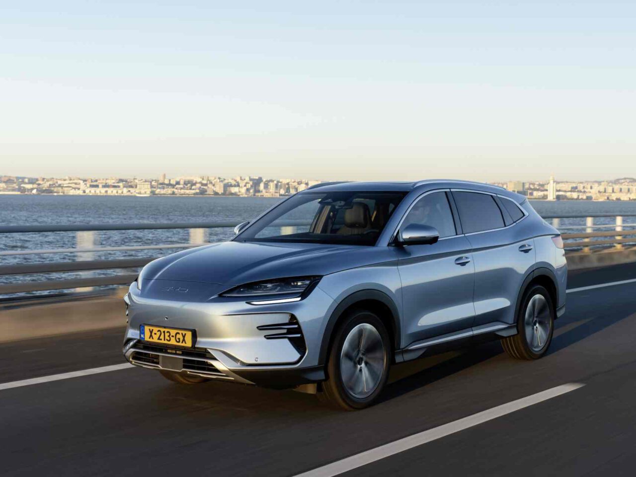Een zilveren BYD Seal U SUV rijdt op een brug aan het water, met de skyline van een stad op de achtergrond.