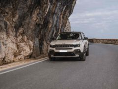 Een zilveren Jeep Avenger rijdt over een bergweg met aan de ene kant een rotsachtige klif en aan de andere kant een slagboom onder een bewolkte hemel.