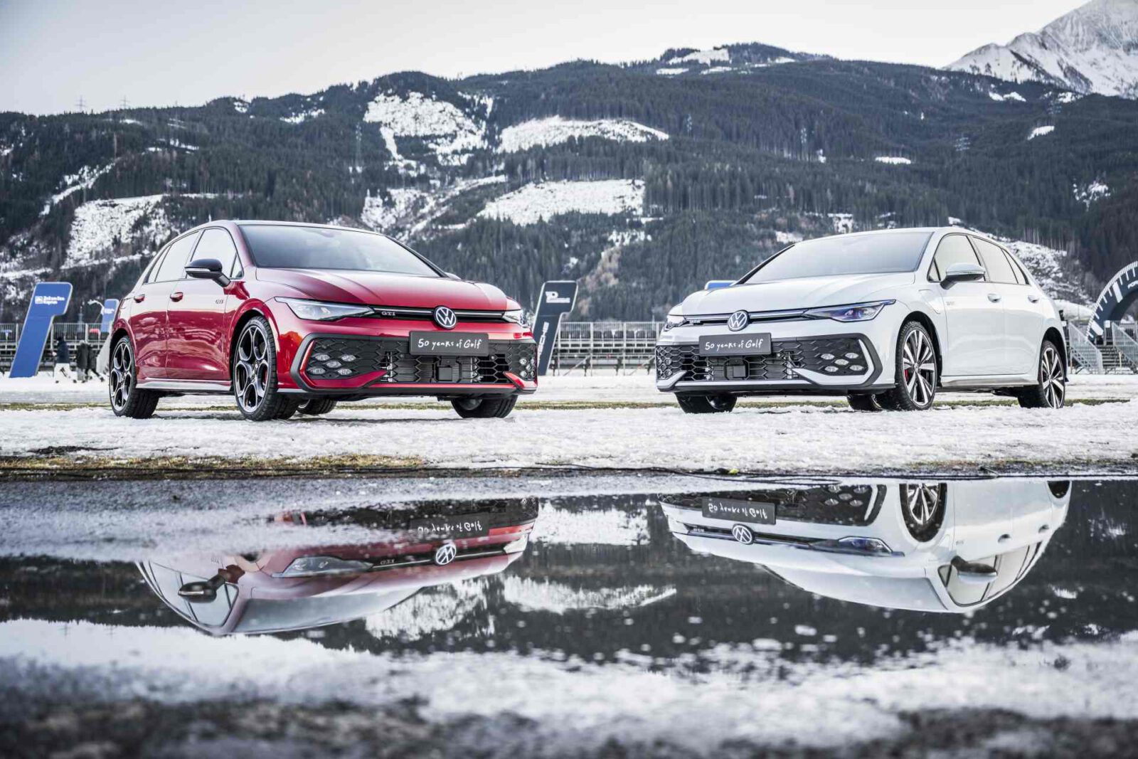Twee Volkswagen Golf GTI-auto's, een rode en een witte, geparkeerd op een besneeuwde grond met een bergachtige achtergrond. Hun reflecties zijn zichtbaar in een nabijgelegen plas.
