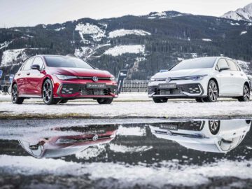 Twee Volkswagen Golf GTI-auto's, een rode en een witte, geparkeerd op een besneeuwde grond met een bergachtige achtergrond. Hun reflecties zijn zichtbaar in een nabijgelegen plas.