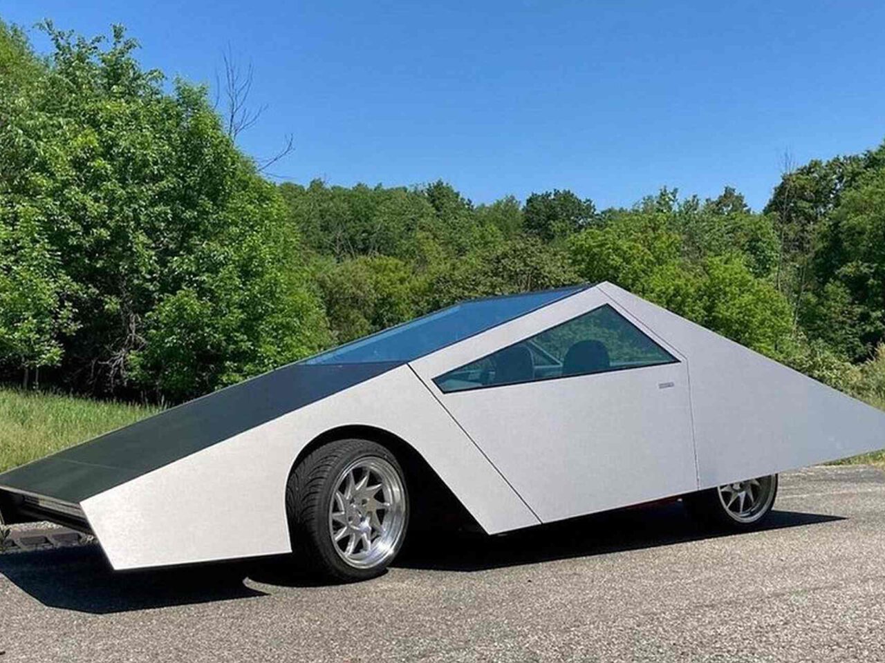 Een futuristische, hoekige cybertruck met een zilver-zwarte buitenkant staat geparkeerd op een verhard oppervlak, omgeven door groen gebladerte en bomen.