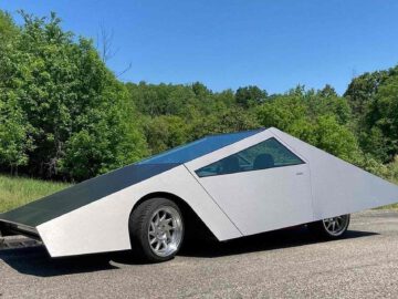 Een futuristische, hoekige cybertruck met een zilver-zwarte buitenkant staat geparkeerd op een verhard oppervlak, omgeven door groen gebladerte en bomen.
