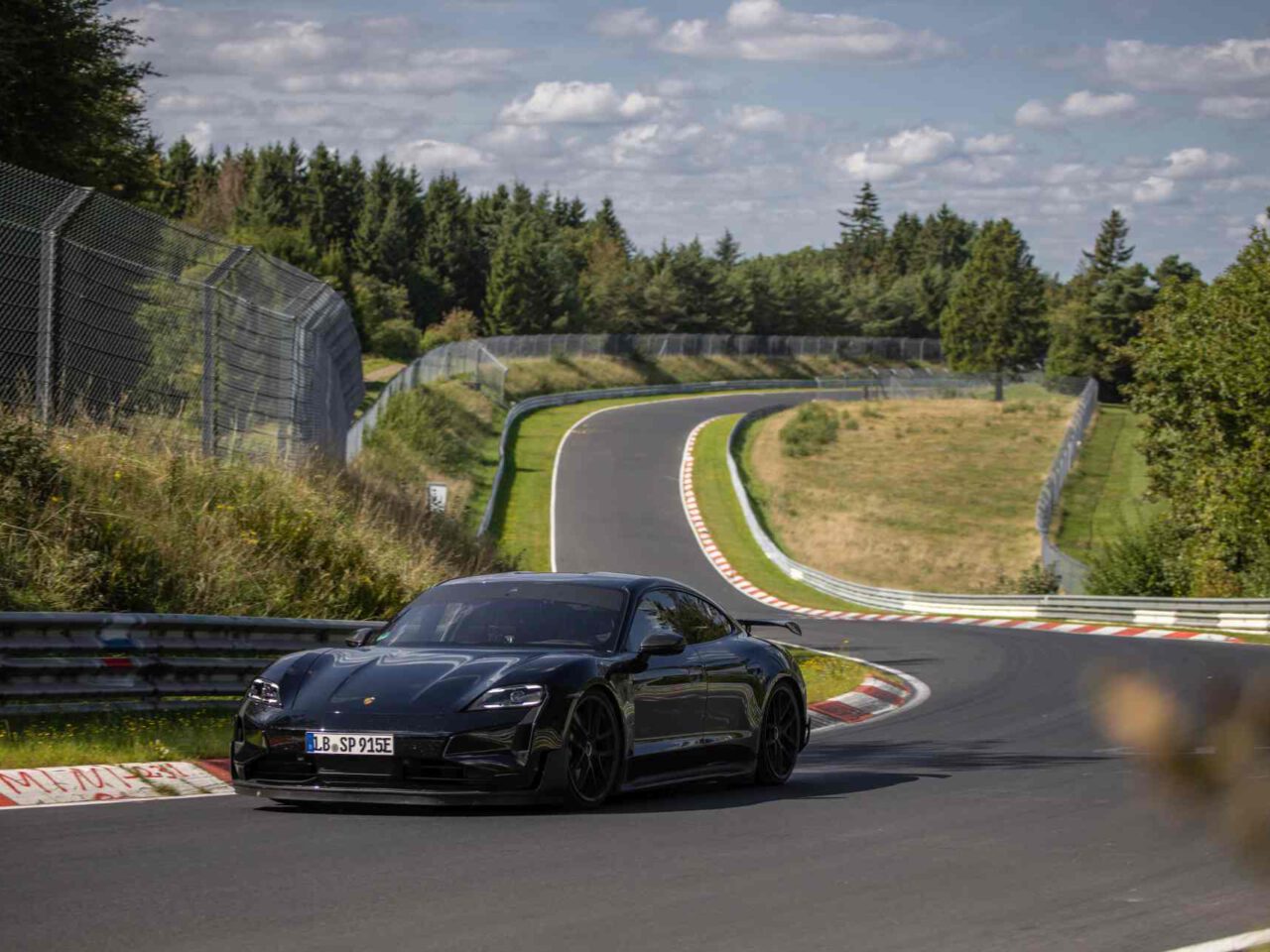 Een strakke zwarte Taycan rijdt op een bochtig racecircuit omgeven door groen en bomen onder een gedeeltelijk bewolkte hemel.