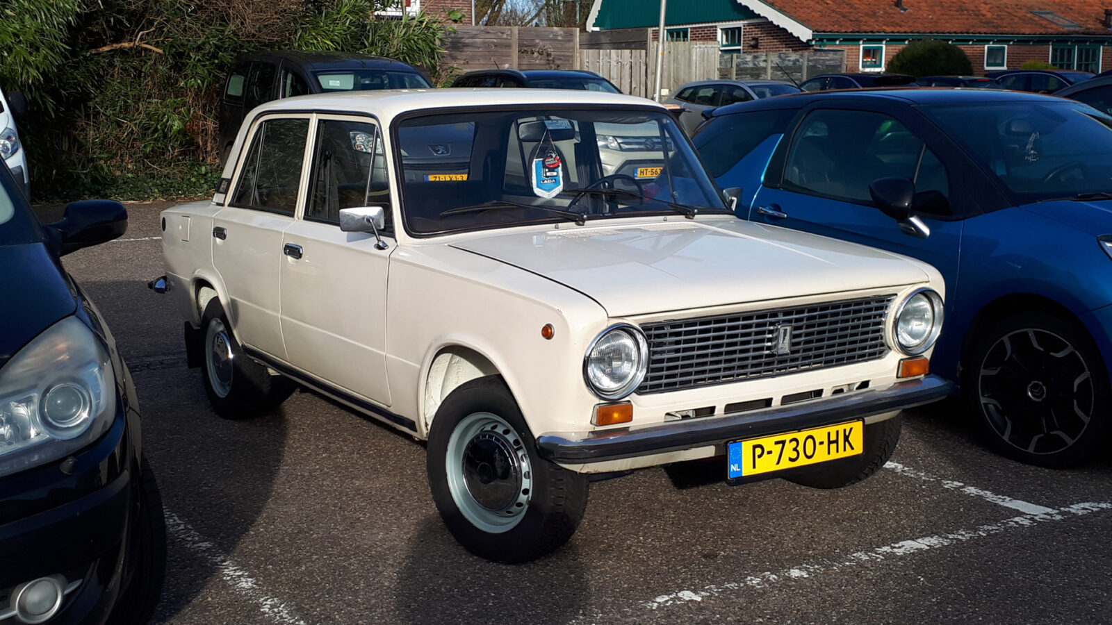 Een vintage witte Lada 1200 geparkeerd op een parkeerplaats, omringd door moderne voertuigen, op een zonnige dag.