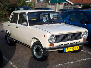 Een vintage witte Lada 1200 geparkeerd op een parkeerplaats, omringd door moderne voertuigen, op een zonnige dag.