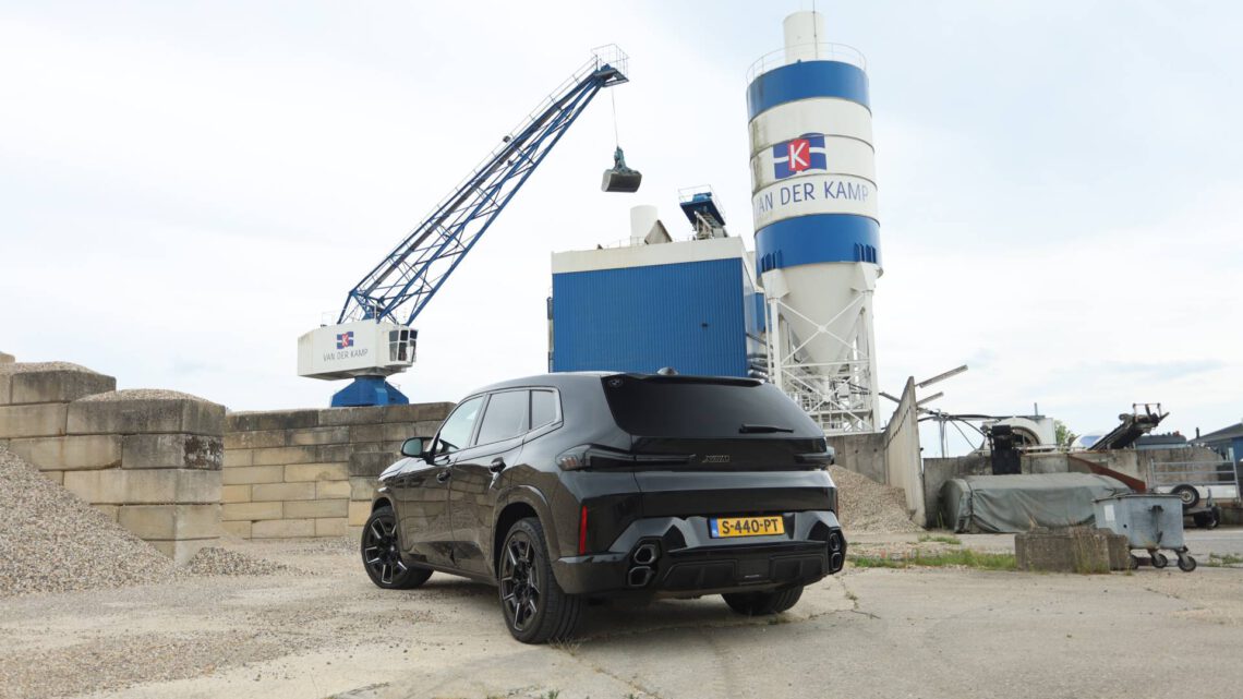 Un BMW XM negro aparcado cerca de una grúa y un gran silo de cemento azul y blanco en un polígono industrial.