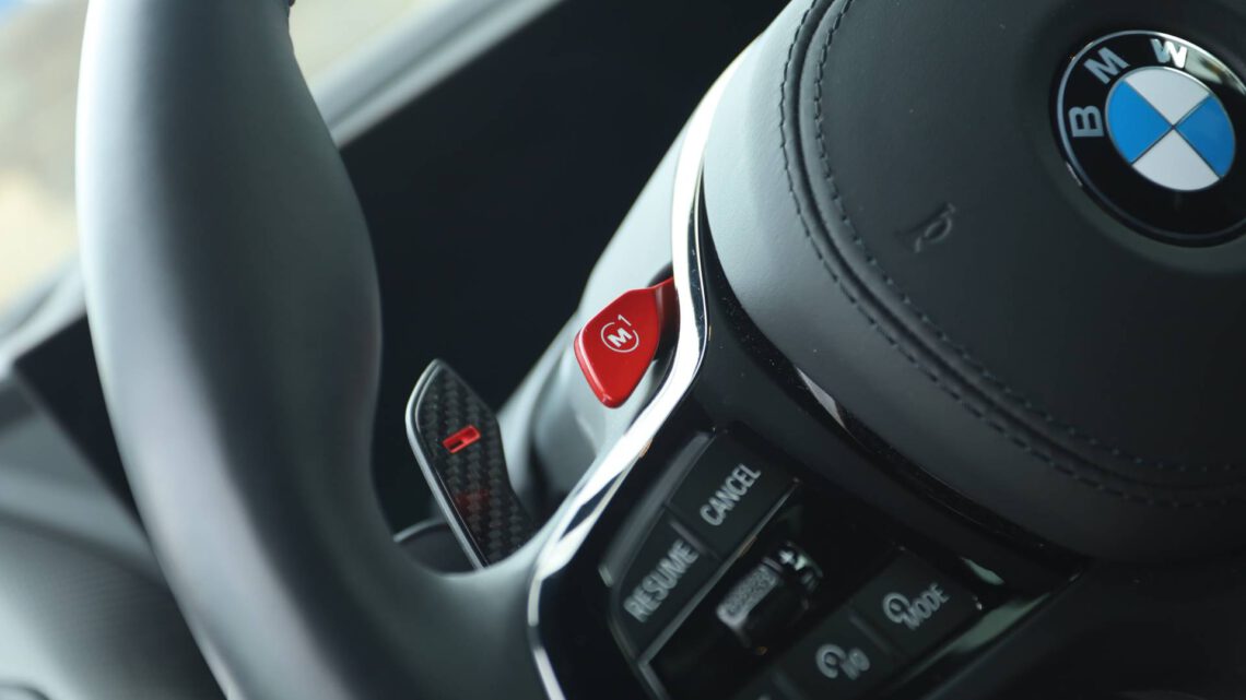 Close-up van een BMW XM-stuur met bedieningselementen en logo, gericht op de rode M1-knop aan de linkerkant.