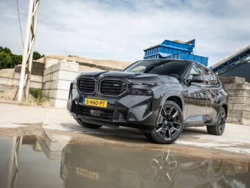 Een BMW XM geparkeerd bij een waterplas op een industriegebied, met betonblokken en een blauwe container op de achtergrond.