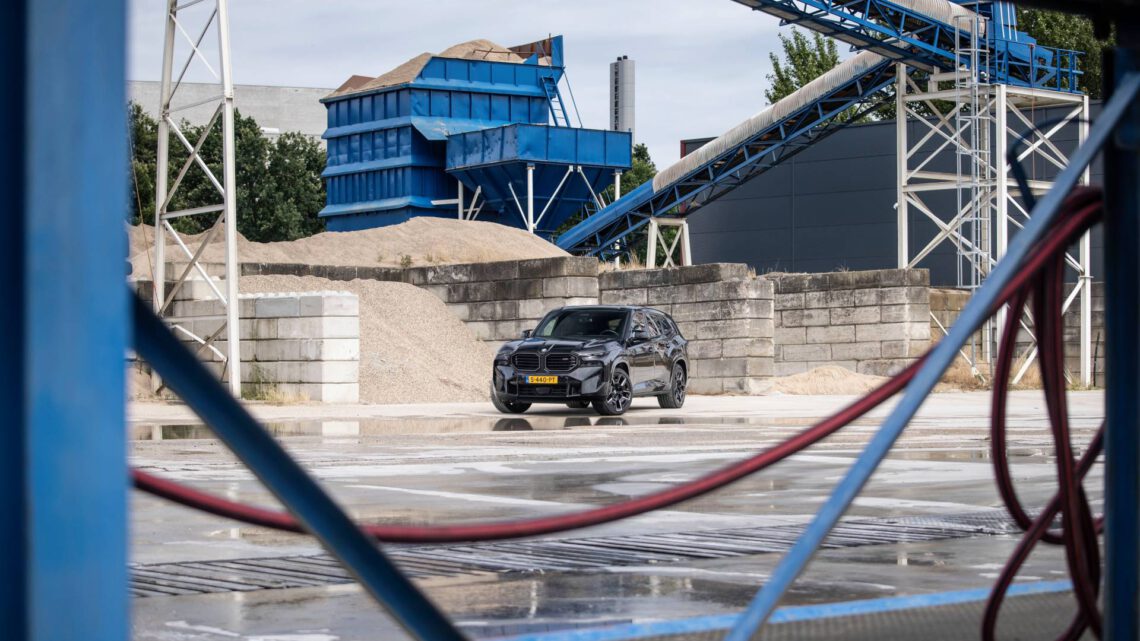 Black BMW XM at an industrial washing station with blue machine structures and hoses in the foreground.