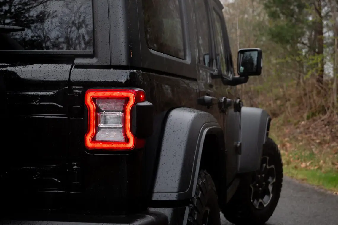 Primer plano de una luz trasera ardiendo en un Jeep Wrangler 4xe negro con el exterior mojado, lo que sugiere lluvia reciente.