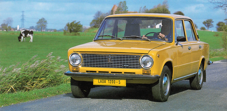 A yellow Lada 1200 car drives through a rural landscape, with grazing cows in the background. A male driver is visible behind the wheel.