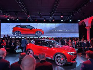 Een rode auto die wordt tentoongesteld op een autoshow met publiek en een presentator ervoor, waarbij de kenmerken van de auto worden benadrukt tegen een verlichte promotionele achtergrond.