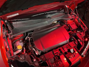 Close-up van een open rode autokap met een gedetailleerd motorcompartiment met zichtbare componenten en branding.