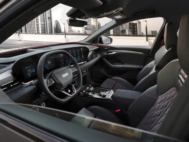 Interior view of an Audi car showing the steering wheel, dashboard and leather seats, with a cityscape visible through the windshield.