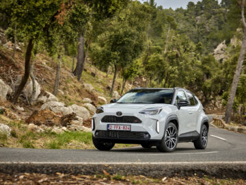 Een witte SUV, mogelijk een Toyota Yaris Cross, rijdt op een bochtige weg door een bosrijk gebied met heuvels en hoge bomen. Op het kenteken staat "2-FFA-824." De scène verwijst naar de 2024-update voor deze Toyota-bestseller.