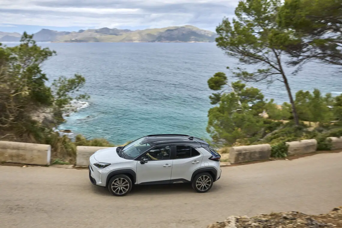 Ein weißer 2024 Toyota Yaris Cross fährt entlang einer Küstenstraße mit atemberaubendem Blick auf das Meer und die Berge im Hintergrund, eingerahmt von üppigen Bäumen.