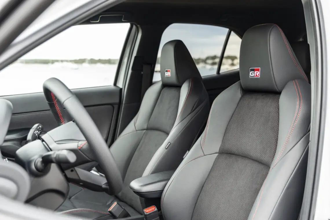 Intérieur des sièges avant de la Toyota Yaris Cross 2024, avec une sellerie grise à surpiqûres rouges, un logo GR sur les dossiers et un tableau de bord épuré avec des éléments de design sportif.