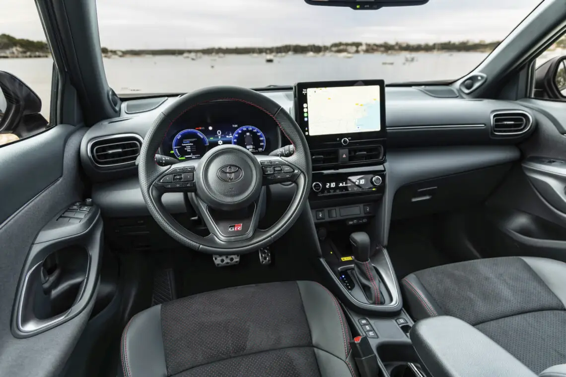 Der Innenraum des Toyota Yaris Cross 2024 bietet ein Lenkrad mit Bedienelementen, ein digitales Armaturenbrett, ein Touchscreen-Display und ein Automatikgetriebe. Draußen können Sie durch die Windschutzscheibe einen Blick auf das Wasser mit Booten werfen. Perfekt für Ihre nächste Autorevue.