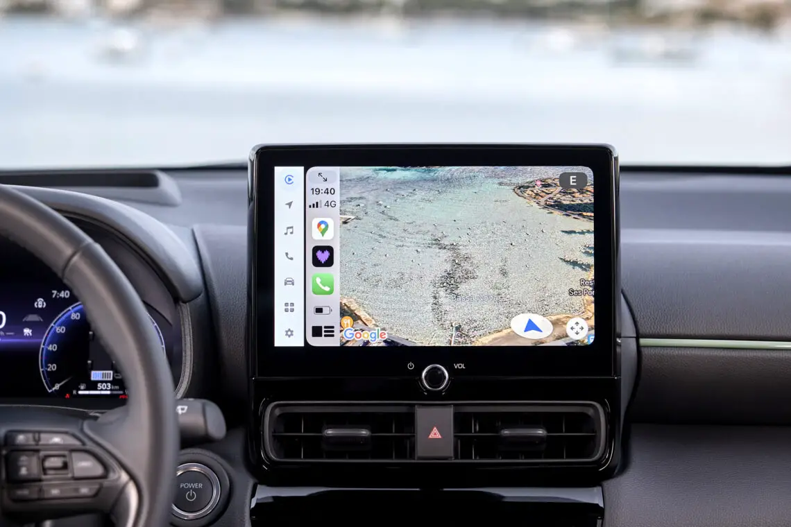 Close-up van het infotainmentsysteem van de Toyota Yaris Cross uit 2024 met navigatie op een kustkaart. Het dashboard en het stuur zijn gedeeltelijk zichtbaar en tonen de nieuwste update op het gebied van autotechnologie.