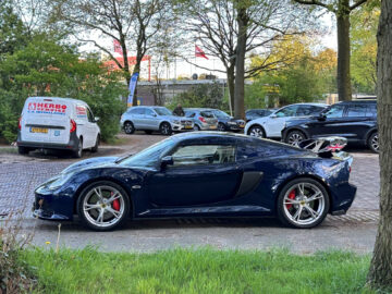 A blue Lotus Exige S parked in a street next to a grassy field, with several other cars and vans nearby under a clear sky.