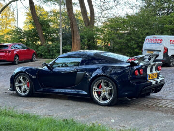A dark blue Lotus Exige S sports car parked in a gravel lot next to green trees, with a white van and a red car in the background.