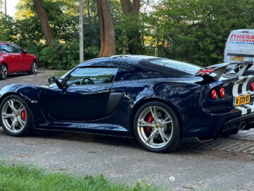 Een zwarte Lotus Exige S geparkeerd op een geplaveide straat vlakbij een park, met andere voertuigen op de achtergrond.